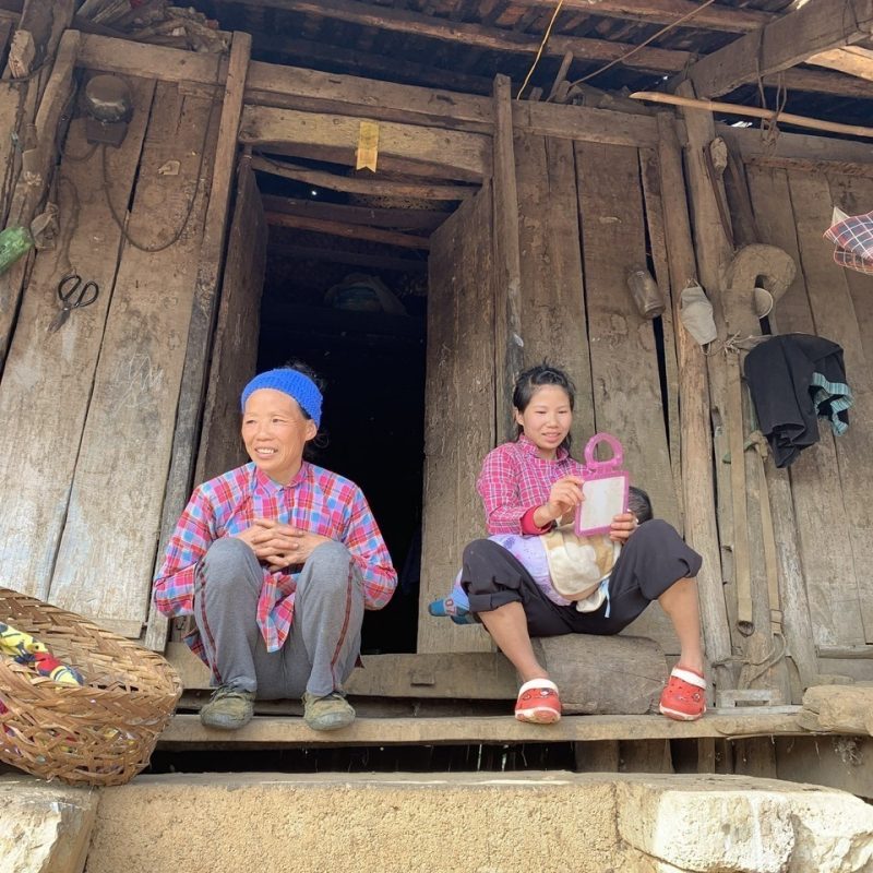 Two women sitting on the steps of a rustic wooden house, smiling and engaging in conversation, with household items and a basket nearby